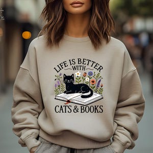 May include: Beige sweatshirt with the text "LIFE IS BETTER WITH CATS & BOOKS" and a graphic of a black cat lying on a book surrounded by flowers. The sweatshirt is a neutral color.