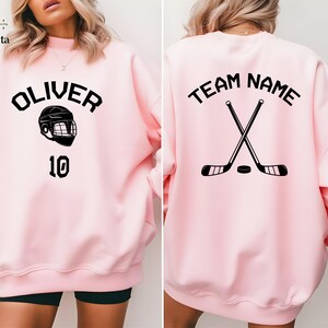 May include: Pink sweatshirt with a hockey helmet and the name "Oliver" and the number "10" on the front. The back of the sweatshirt has the text "Team Name" with two crossed hockey sticks.