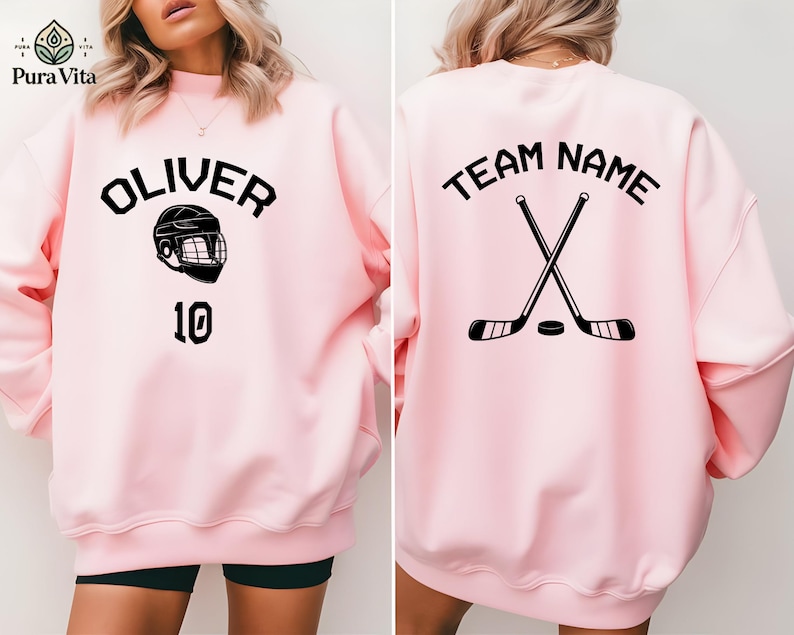 May include: Pink sweatshirt with a hockey helmet and the name "Oliver" and the number "10" on the front. The back of the sweatshirt has the text "Team Name" with two crossed hockey sticks.