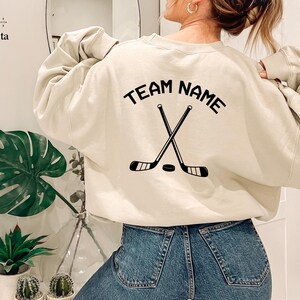 May include: A beige sweatshirt with the text "TEAM NAME" printed on the back. The text is in a black font and is surrounded by a black hockey stick and puck graphic.