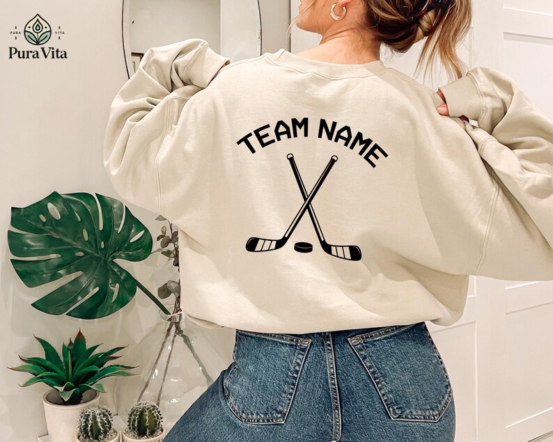 May include: A beige sweatshirt with the text "TEAM NAME" printed on the back. The text is in a black font and is surrounded by a black hockey stick and puck graphic.