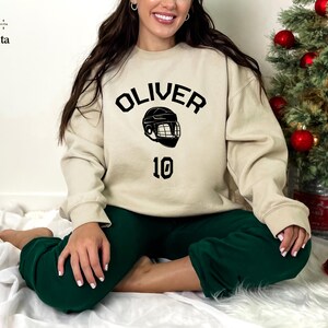 May include: A beige sweatshirt with black text that reads "OLIVER" and a hockey helmet graphic. The number "10" is printed below the graphic.