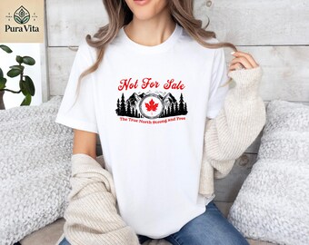 May include: White t-shirt with a graphic design featuring a red maple leaf, mountains, and the text "Not For Sale" and "The True North Strong and Free". The design is in red and black.