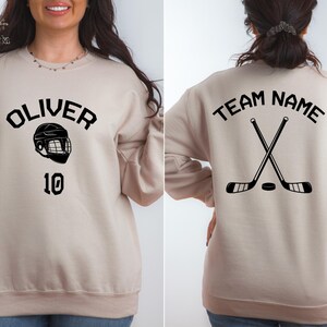 May include: Two beige sweatshirts with hockey-themed designs. One sweatshirt has the text "OLIVER" and a hockey helmet graphic with the number "10" below. The other sweatshirt has the text "TEAM NAME" and a graphic of two crossed hockey sticks with a puck.