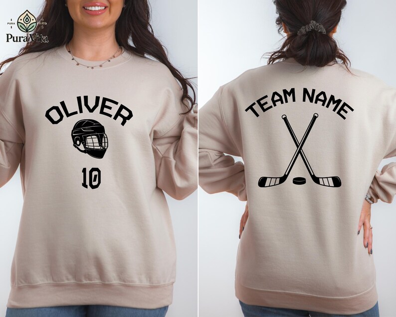 May include: Two beige sweatshirts with hockey-themed designs. One sweatshirt has the text "OLIVER" and a hockey helmet graphic with the number "10" below. The other sweatshirt has the text "TEAM NAME" and a graphic of two crossed hockey sticks with a puck.