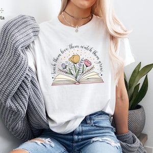 May include: White t-shirt featuring a colorful illustration of an open book with flowers and the text "Teach them love them watch them grow." The design includes yellow, pink, and green floral elements, perfect for teachers or book lovers.