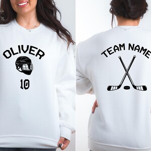 May include: White sweatshirt with black text and graphic. The front of the sweatshirt says "OLIVER" with a hockey helmet graphic and the number "10". The back of the sweatshirt says "TEAM NAME" with two hockey sticks crossed over each other.