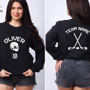 May include: Black sweatshirt with white text and graphic. The text on the front says "Oliver" and has a hockey helmet graphic with the number "10" below it. The back of the sweatshirt says "Team Name" with two crossed hockey sticks and a puck.