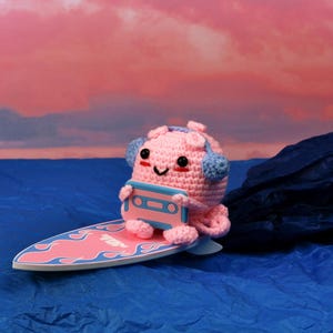 May include: A crocheted pink octopus wearing blue headphones and holding a cassette tape, riding a pink and blue surfboard. The background features a blue ocean and a pink and orange sky.