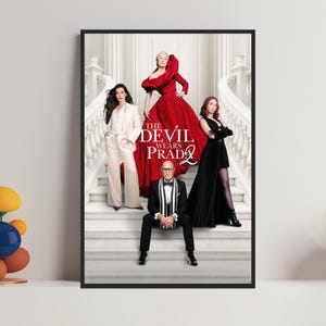 Puede incluir: Un póster enmarcado de la película "El diablo viste de Prada 2" con cuatro personas en una gran escalera. El póster tiene un marco negro y el título se muestra de forma destacada en texto blanco y rojo.