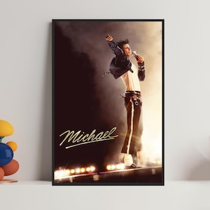 Puede incluir: Un póster enmarcado con Michael Jackson en el escenario. Lleva una chaqueta negra, una camisa blanca y pantalones negros con una raya dorada. La palabra "Michael" está escrita en letras doradas.