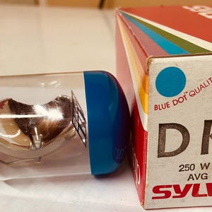 May include: A clear glass light bulb with a copper base and a blue cap. The bulb is labeled "DKM 250 W - 21.5 V AVG 25 HRS SYLVANIA" and "BLUE DOT QUALITY LAMPS".