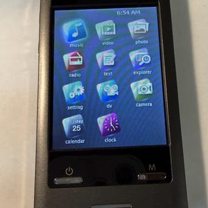 May include: A dark gray portable media player with a touch screen displaying app icons for music, video, photos, and more. The screen shows the time as 6:54 AM and the date as Friday, the 25th.