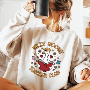 May include: A light beige sweatshirt with a graphic of a goose wearing a cap and reading a book. The graphic is surrounded by hearts, stars, and flowers. The text "Silly Goose Teacher Club" is printed in a circle around the graphic.