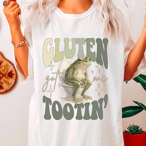 May include: White t-shirt with a humorous graphic. The design features a frog sitting on a toilet with the text "GLUTEN got me TOOTIN'" in green and beige lettering. The shirt has a relaxed fit.