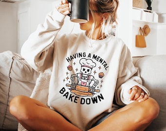 May include: A beige crewneck sweatshirt featuring a graphic of a skeleton wearing a chef hat and apron, using a rolling pin on dough. The text reads HAVING A MENTAL BAKE DOWN, accompanied by small illustrations of a strawberry and a muffin.