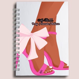 Peut inclure: Un agenda à spirales avec une couverture représentant une illustration d'une paire de jambes portant des talons hauts roses avec un nœud. Le texte "My Planner 2025" et "Pray | Execute | Believe" figurent également sur la couverture.