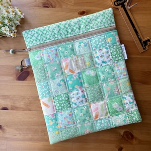 May include: A green patchwork zippered case with a variety of floral and patterned fabrics. The case has a light brown zip and a small tag that reads "Little Jamie". A small bunch of white flowers and a wooden ruler are in the background.