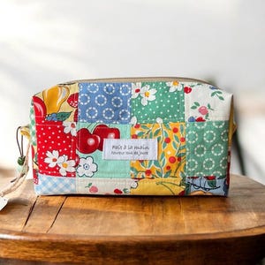 May include: A rectangular fabric pouch featuring a patchwork design of various colorful patterns including cherries, flowers, and fruits. A white rectangular label on the front displays the text Fait à la main heureux tous les jours.