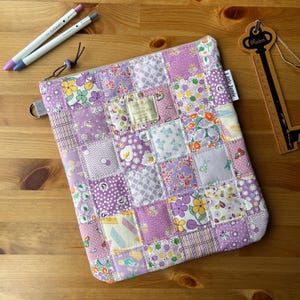 May include: A patchwork zipped pouch in various shades of purple, with floral and geometric designs. The pouch has a metal ring and a tag that reads "Little Jamie". Two pens are visible in the upper left corner.