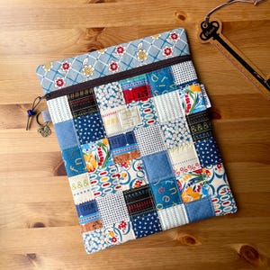 May include: A rectangular fabric pouch featuring a patchwork design of various blue, white, red, and yellow patterned squares. The top section has a blue floral print with a brown zipper closure. A small tag reads Little Jamie.