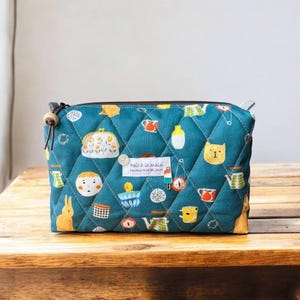 Peut inclure: Pochette matelassée turquoise avec fermeture éclair et tirette en perle de bois. Décorée d'illustrations de dessins animés, dont un chat, un lapin et des tasses. Le texte "Fait à la main" est imprimé sur une étiquette blanche.