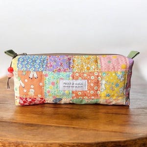 May include: A rectangular quilted pencil case featuring a colourful patchwork design of various floral and animal patterns in shades of yellow, orange, pink, and blue. A white fabric label reads Fait à la main, heureux tous les jours.