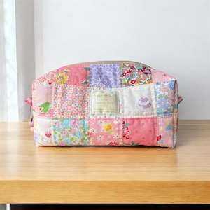 May include: A rectangular fabric pouch featuring a patchwork design of various floral and patterned cotton squares in shades of pink, purple, blue, and white. A central rectangular label reads It's Lovely Day. The pouch has a top zipper closure.