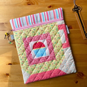 May include: A quilted fabric pouch featuring a central square with a red and blue cupcake graphic, surrounded by yellow floral and pink polka dot patterns. The top has a pink zip and a multi-coloured striped border. A key-shaped ruler is visible.