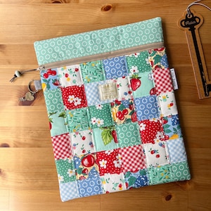 May include: A rectangular zipped pouch featuring a patchwork design of various fabric squares in red, green, blue, and white. The patterns include strawberries, cherries, gingham, and floral motifs. A small metal tag is attached to the zip pull.