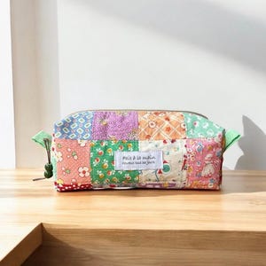 May include: A rectangular zipped pouch made of patchwork fabric featuring various floral and geometric patterns in shades of pink, green, orange, and purple. A white rectangular label on the front displays the text Fait à la main heureux tous les jours.