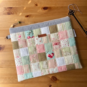 May include: A rectangular zipped pouch featuring a patchwork design of various floral and cherry-patterned fabric squares in shades of pink, green, and white. The pouch has a grey fabric top border, a brown leather label, and a small green bead zip pull.