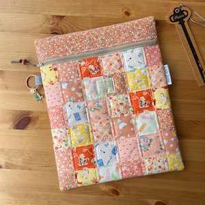 May include: A rectangular zippered pouch featuring a patchwork design of orange, yellow, and white fabric squares with various cartoon illustrations. The pouch includes a small metal key-shaped charm attached to the zipper pull and a white tag labeled Little Jamie.