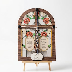 Puede incluir: Una obra de arte en vitral con marco de madera, con un diseño floral y texto. La obra de arte presenta una cuerda trenzada y una pequeña etiqueta con nombres y una fecha. La pieza se exhibe en un pequeño caballete de madera.