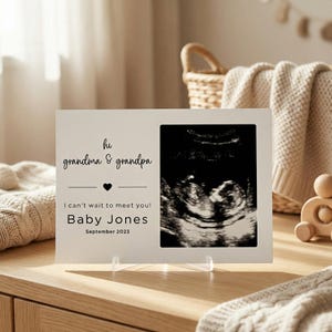 Op de afbeelding: Een witte fotolijst met een echo en de tekst "hi grandma & grandpa", een hartsymbool en "I can't wait to meet you! Baby Jones September 2023". De lijst staat op een transparante standaard.