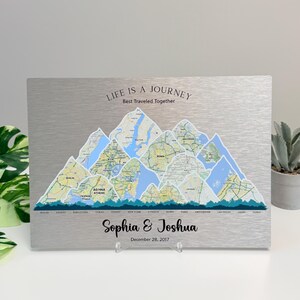 Puede incluir: Un letrero de metal plateado con un diseño de montaña con detalles de mapas. El letrero incluye el texto "LIFE IS A JOURNEY Best Traveled Together", los nombres "Sophia & Joshua" y la fecha "December 28, 2017".