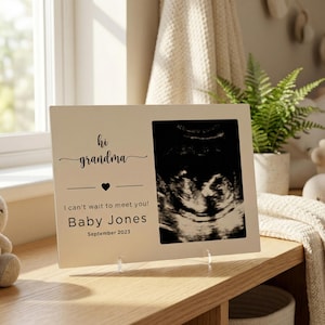 Puede incluir: Un marco de fotos color crema con el texto "hi grandma" y "I can't wait to meet you! Baby Jones September 2023." El marco incluye una imagen de ultrasonido y está apoyado sobre una superficie de madera.