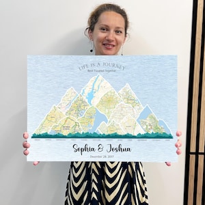 Puede incluir: Una impresión en lienzo azul claro con un diseño de cordillera compuesto por mapas de ciudades. El texto "LIFE IS A JOURNEY Best Traveled Together" está en la parte superior. Debajo, los nombres "Sophia & Joshua" y la fecha "December 28, 2017" se muestran.