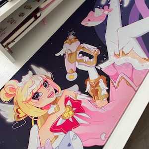 May include: A rectangular desk mat featuring a vibrant anime-style illustration of a character with blonde hair, wearing a pink and white outfit with heart accents, holding a pink toy gun, and surrounded by small shark figures on a dark background.