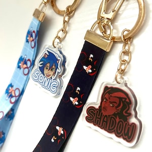 May include: Close-up of three lanyards with gold-colored hardware. One lanyard is light blue with a repeating Sonic the Hedgehog pattern. The second is navy blue with a repeating Shadow the Hedgehog pattern. The third has a Shadow the Hedgehog charm with the name "SHADOW".