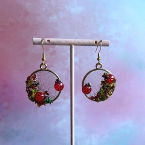 May include: A pair of circular gold-toned earrings featuring clear resin centers embedded with green moss, small red ladybug charms, and fine gold glitter. The earrings hang from gold-toned fishhook wires and are displayed on a stand.
