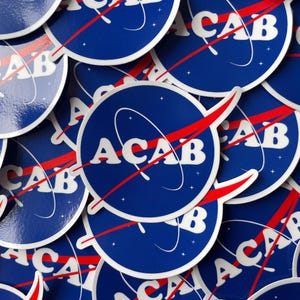 May include: A collection of circular stickers featuring a dark blue background with white stars and the text ACAB in a bold white font. A red swoosh graphic crosses the center of each sticker, mimicking a space agency logo design.