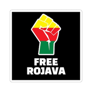 May include: A square sticker with a black background features a raised fist in red, yellow, and green. The words "FREE ROJAVA" are printed in white block letters below the fist. The sticker has a white border.