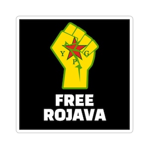 May include: A sticker with a black background featuring a yellow fist with a red star and the letters YPG. Below the fist, the words "FREE ROJAVA" are printed in white.