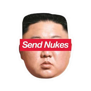 Può includere: Un adesivo con un ritratto e uno striscione rosso sugli occhi. Lo striscione recita "Send Nukes" in lettere maiuscole bianche. Il ritratto è di un uomo con capelli corti neri e un'espressione neutra.