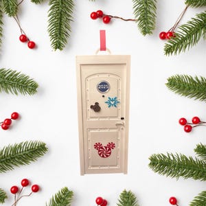 May include: A cream-colored door-shaped Christmas ornament with a red ribbon for hanging. The door has a blue circle with "2025" and Mickey Mouse, snowflake, and candy cane designs. The ornament is surrounded by evergreen branches and red berries.