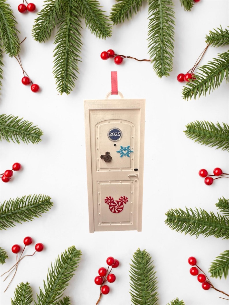 May include: A cream-colored door-shaped Christmas ornament with a red ribbon for hanging. The door has a blue circle with "2025" and Mickey Mouse, snowflake, and candy cane designs. The ornament is surrounded by evergreen branches and red berries.