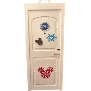 May include: White rectangular ornament with a door design. Features a Mickey Mouse head made of candy canes, a snowflake, and a Mickey Mouse head. The year 2025 is displayed on the top panel. A pink ribbon is attached for hanging.