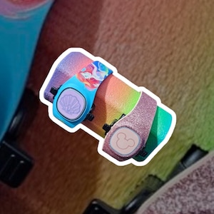 May include: Two colorful Disney MagicBands with a rainbow design. One band features a blue shell with a mermaid design and the other band features a pink Mickey Mouse head.