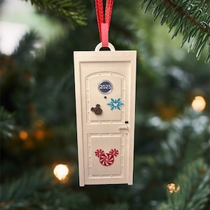 May include: A white rectangular Christmas ornament shaped like a door, with a red ribbon for hanging. The door features a Mickey Mouse candy cane design, a snowflake, and the year 2025. The ornament hangs in front of a blurred Christmas tree.